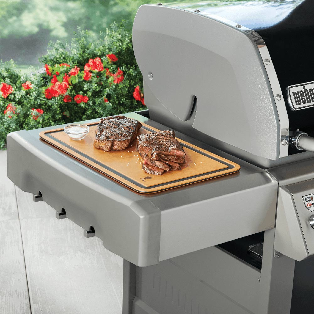 Cutting board with tan surface and dark border, resting on a grill surface with food portions.