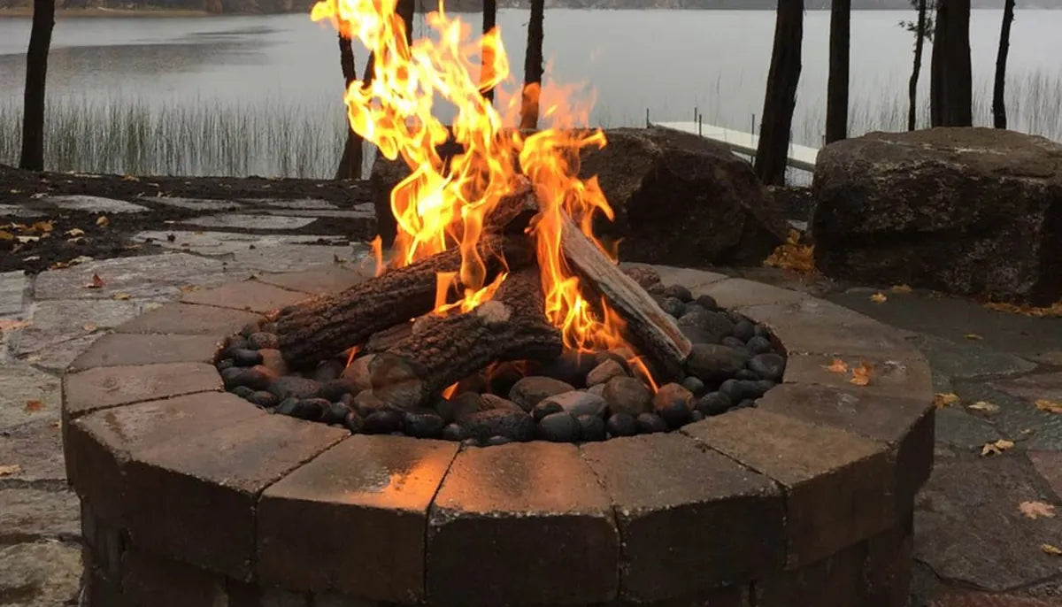 Round stone gas fire pit with lava rock and ceramic log set burning near a lakeside patio — gas supply and BTU performance