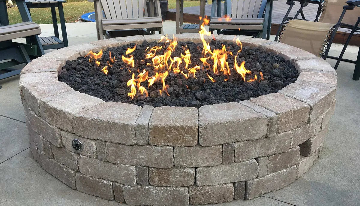 Large round concrete block fire pit with lava rock media burning on a backyard concrete patio surrounded by Adirondack chairs