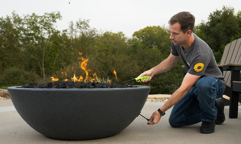 Spotix team member lighting a large dark concrete fire pit bowl with lava rocks on a patio surrounded by trees